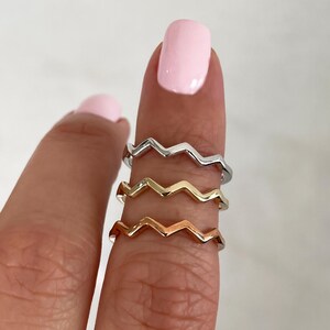 Pode incluir: Tr&ecirc;s an&eacute;is de metal finos empilhados num dedo, cada um com um padr&atilde;o distinto de ziguezague ou chevron. O anel superior &eacute; prateado, o do meio &eacute; dourado e o inferior &eacute; de ouro rosa, todos com um acabamento liso e polido.