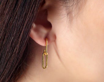 May include: A gold-coloured, paperclip-shaped earring is shown. The earring is a long, rectangular shape with a smaller rectangle attached at the top. The earring is hanging from the ear of a person with dark hair.