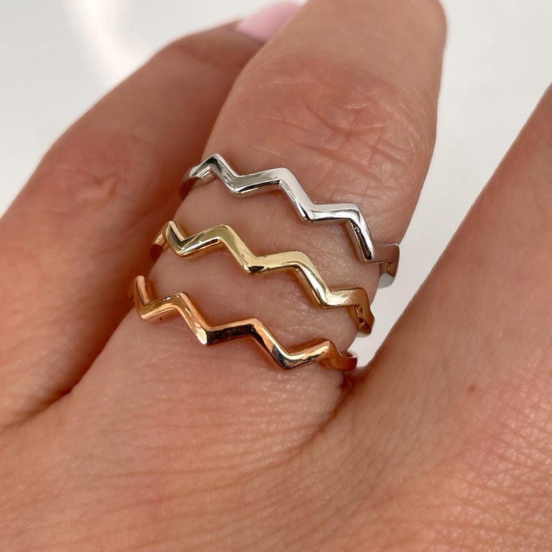 Pode incluir: Um conjunto de tr&ecirc;s an&eacute;is de metal finos com um padr&atilde;o repetitivo de ziguezague ou chevron. Os an&eacute;is t&ecirc;m tr&ecirc;s tons met&aacute;licos distintos: prata brilhante, ouro amarelo quente e ouro rosa suave, empilhados juntos em um dedo.