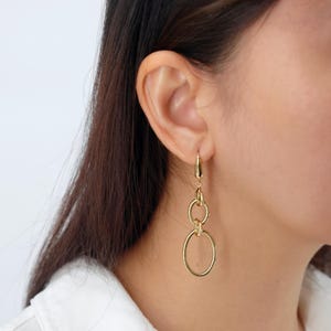 May include: Gold-toned dangle earrings featuring a series of connected oval and circular links. The earrings are suspended from a small hoop that attaches to the ear. The design is simple and elegant, with a polished finish.