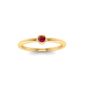 May include: A gold ring with a small, round, red gemstone set in the center. The band is thin and polished, and the gemstone is a vibrant red color. The ring is set against a white background.