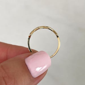 Pode incluir: Um anel fino de ouro com uma s&eacute;rie de pequenos segmentos retangulares de topo plano ao longo da parte superior da banda. O metal tem um acabamento polido e reflexivo, com uma estrutura circular cont&iacute;nua e delgada.