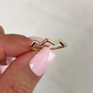 Pode incluir: Um anel de ouro com um padr&atilde;o cont&iacute;nuo de ziguezague ou chevron ao longo da banda superior. O metal tem um acabamento polido e reflexivo, e o design consiste em tr&ecirc;s picos e dois vales distintos, criando uma est&eacute;tica geom&eacute;trica.