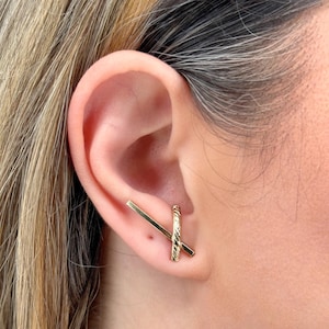 May include: A pair of gold ear cuffs. One cuff is a straight bar and the other is a twisted, curved bar. The cuffs are worn in the upper ear.