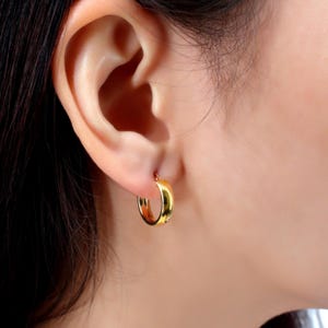 May include: A close-up of a gold hoop earring worn on an ear. The earring is a thick, rounded design, reflecting light with a shiny finish. The skin tone is fair, and the hair is dark.