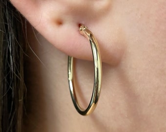 May include: A polished gold hoop earring with a smooth, rounded surface. The circular earring features a hinged closure mechanism at the top where it connects to the earlobe, showcasing a simple, continuous design with a reflective metallic finish.
