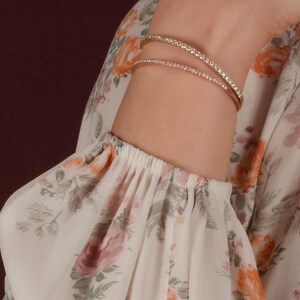 Puede incluir: Dos pulseras doradas, cada una adornada con peque&ntilde;as piedras brillantes, se llevan en la mu&ntilde;eca. Las pulseras destacan sobre una manga estampada con flores en tonos blancos, rosas y naranjas. Se aprecia la mu&ntilde;eca.