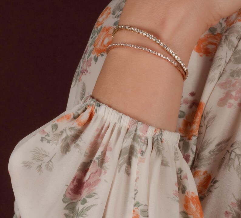 Puede incluir: Dos pulseras doradas, cada una adornada con peque&ntilde;as piedras brillantes, se llevan en la mu&ntilde;eca. Las pulseras destacan sobre una manga estampada con flores en tonos blancos, rosas y naranjas. Se aprecia la mu&ntilde;eca.