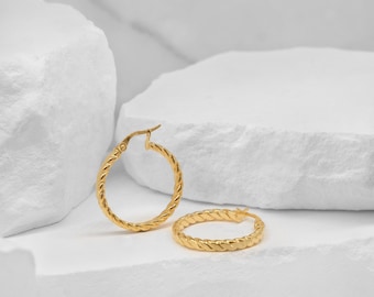 May include: Two gold-colored hoop earrings with a twisted rope design. The earrings are displayed on a white, textured surface. The earrings are a classic jewelry item.