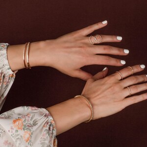 Puede incluir: Juego de joyas doradas sobre un fondo oscuro. La imagen presenta dos pulseras, un anillo infinito y otros tres anillos. Las pulseras y los anillos est&aacute;n adornados con peque&ntilde;as piedras brillantes. Las manos tienen esmalte de u&ntilde;as blanco.