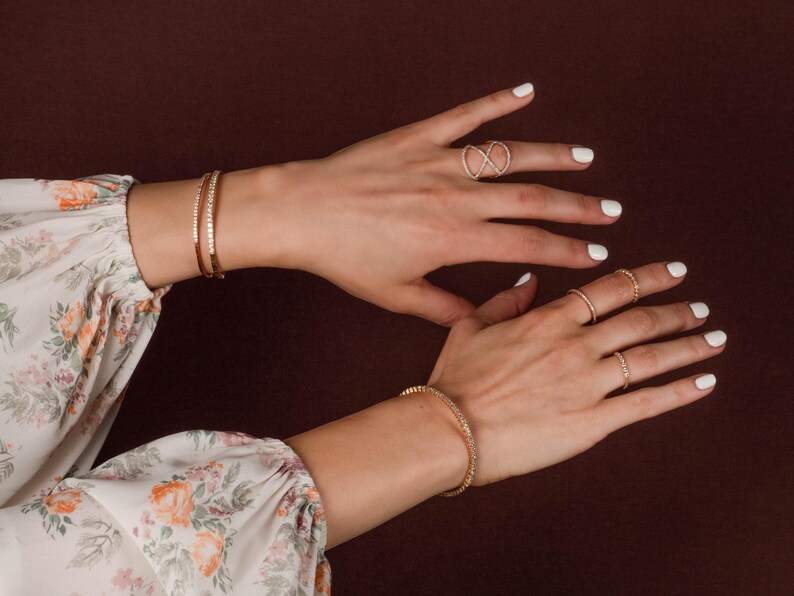 Puede incluir: Juego de joyas doradas sobre un fondo oscuro. La imagen presenta dos pulseras, un anillo infinito y otros tres anillos. Las pulseras y los anillos est&aacute;n adornados con peque&ntilde;as piedras brillantes. Las manos tienen esmalte de u&ntilde;as blanco.