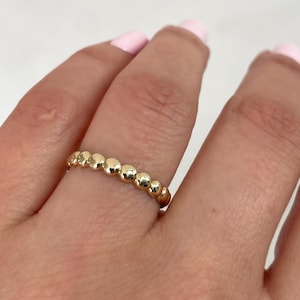 May include: A gold ring featuring a continuous band of small, polished, spherical beads arranged in a row. The metal has a bright, reflective finish, creating a uniform, textured appearance around the entire circumference of the circular jewelry piece.