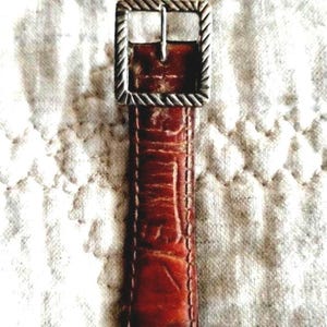 May include: Close-up of a brown leather belt with a silver-colored buckle. The belt has a textured surface and a rectangular buckle with a rope-like design around the edges. The belt is approximately 1 inch wide.