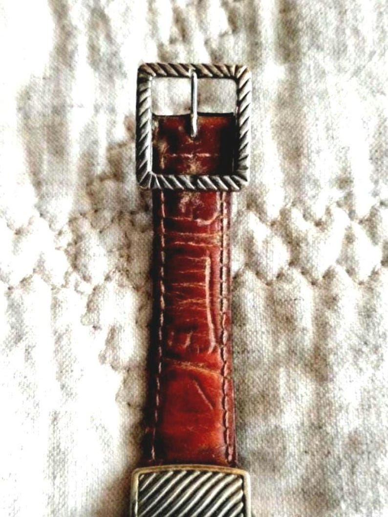 May include: Close-up of a brown leather belt with a silver-colored buckle. The belt has a textured surface and a rectangular buckle with a rope-like design around the edges. The belt is approximately 1 inch wide.