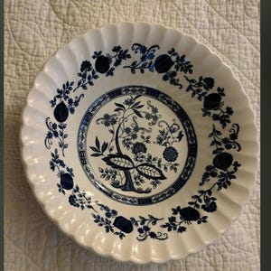 Op de afbeelding: Een witte keramische plaat met een blauw bloemmotief. De plaat heeft een geschulpte rand en een gedetailleerd patroon van bloemen en bladeren in het midden en rond de rand. De plaat is rond.