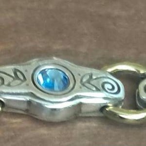May include: A close-up view of a silver and gold bracelet. Each silver link is adorned with a blue gemstone and decorative swirls. The bracelet's links are connected by gold-coloured connectors, creating a striking contrast.