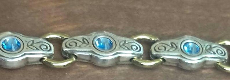 May include: A close-up view of a silver and gold bracelet. Each silver link is adorned with a blue gemstone and decorative swirls. The bracelet's links are connected by gold-coloured connectors, creating a striking contrast.