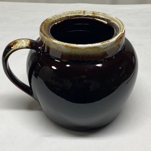 Puede incluir: Un tarro de cerámica marrón con tapa y asa. El tarro tiene un borde blanco y un esmalte marrón.