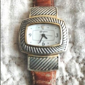 May include: A rectangular watch with a white face, gold trim, and silver-colored, textured accents. The watch has a brown leather band. The watch face displays the brand name and the numbers 3, 6, and 9.