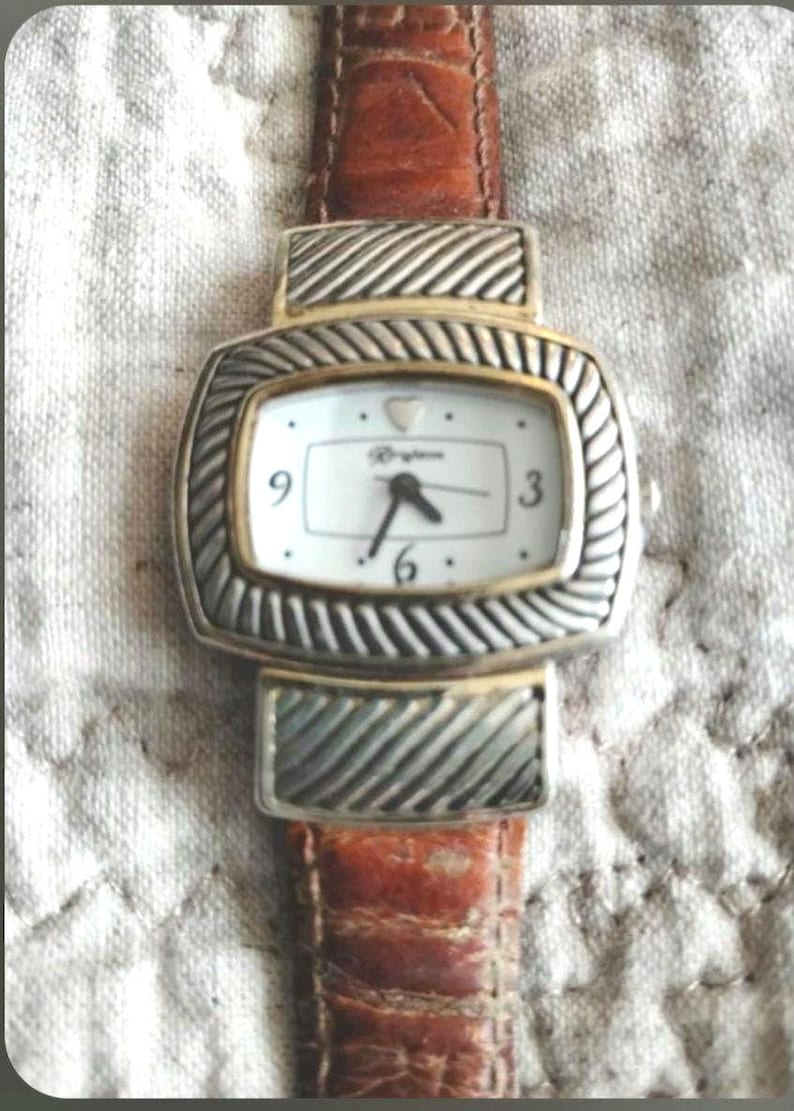 May include: A rectangular watch with a white face, gold trim, and silver-colored, textured accents. The watch has a brown leather band. The watch face displays the brand name and the numbers 3, 6, and 9.