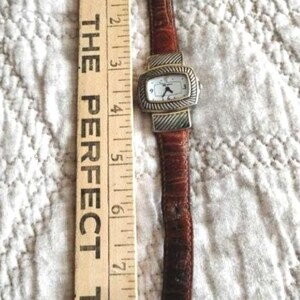 May include: A rectangular watch with a white face and silver-colored bezel, paired with a brown leather band. The watch is next to a wooden ruler with the text "THE PERFECT TOUCH".