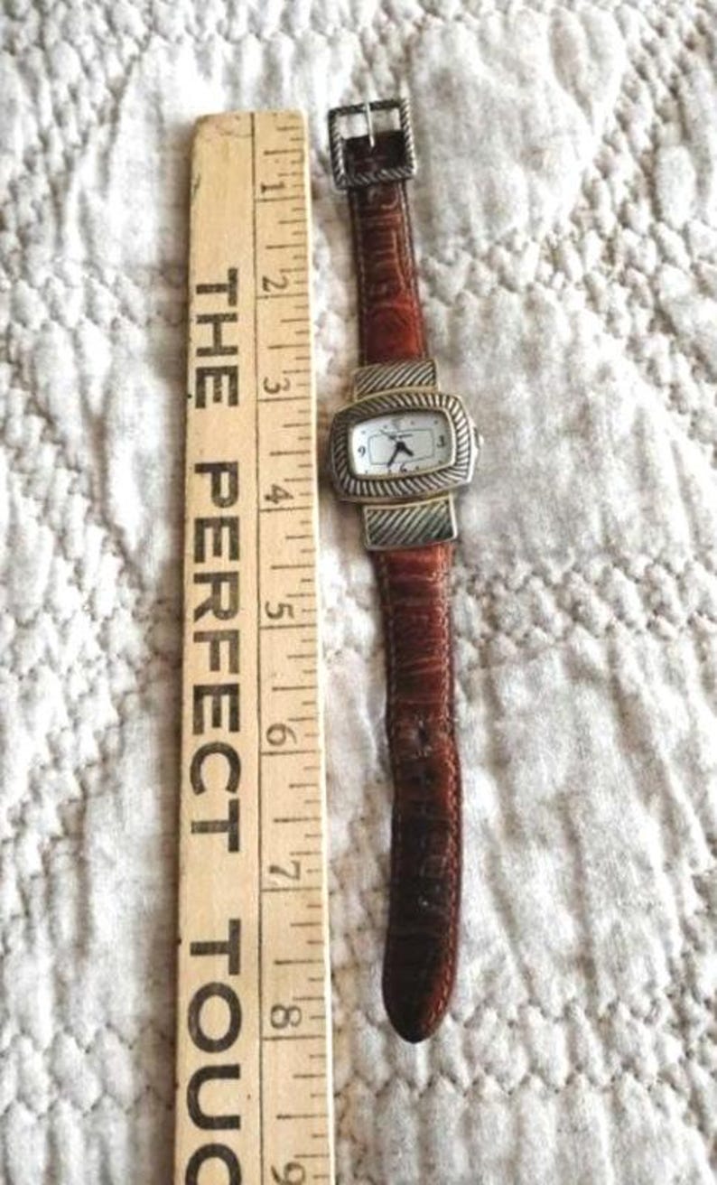 May include: A rectangular watch with a white face and silver-colored bezel, paired with a brown leather band. The watch is next to a wooden ruler with the text "THE PERFECT TOUCH".