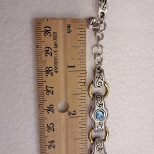 May include: Silver bracelet with gold details and blue gemstones. The bracelet is measured with a ruler, showing a length of approximately 7.6 cm.