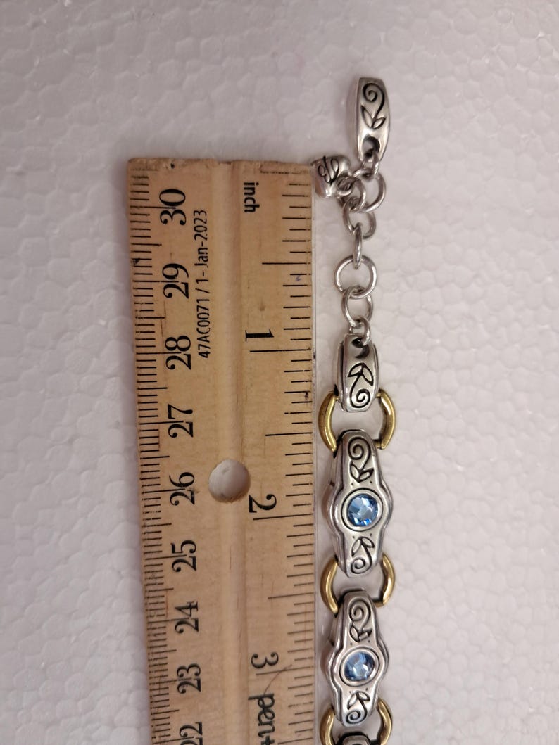 May include: Silver bracelet with gold details and blue gemstones. The bracelet is measured with a ruler, showing a length of approximately 7.6 cm.