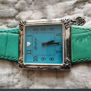 May include: A square-faced watch with a turquoise dial and silver-tone case. The watch has a turquoise leather band. The watch face displays the brand name "Brighton". The watch hands point to 7:00. The watch is on a textured, gray surface.