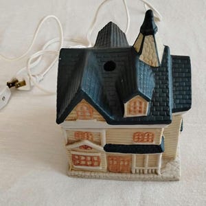 May include: A ceramic, illuminated miniature house with a blue roof, beige walls, and brown trim. The house has multiple windows and a chimney. A white electrical cord and plug are attached.