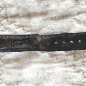 May include: Close-up of a black leather watch strap with a silver buckle. The strap has several holes for adjustment and is stitched along the edges. The leather appears worn, with visible creases and imperfections.