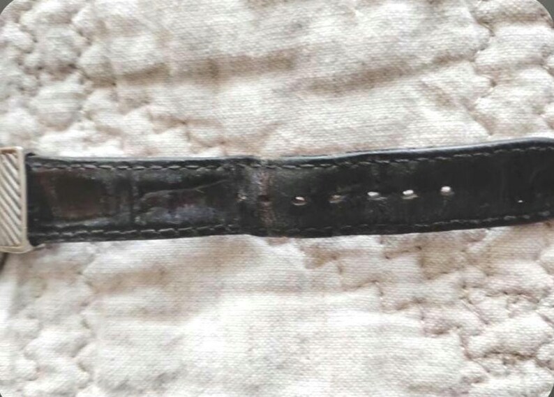 May include: Close-up of a black leather watch strap with a silver buckle. The strap has several holes for adjustment and is stitched along the edges. The leather appears worn, with visible creases and imperfections.