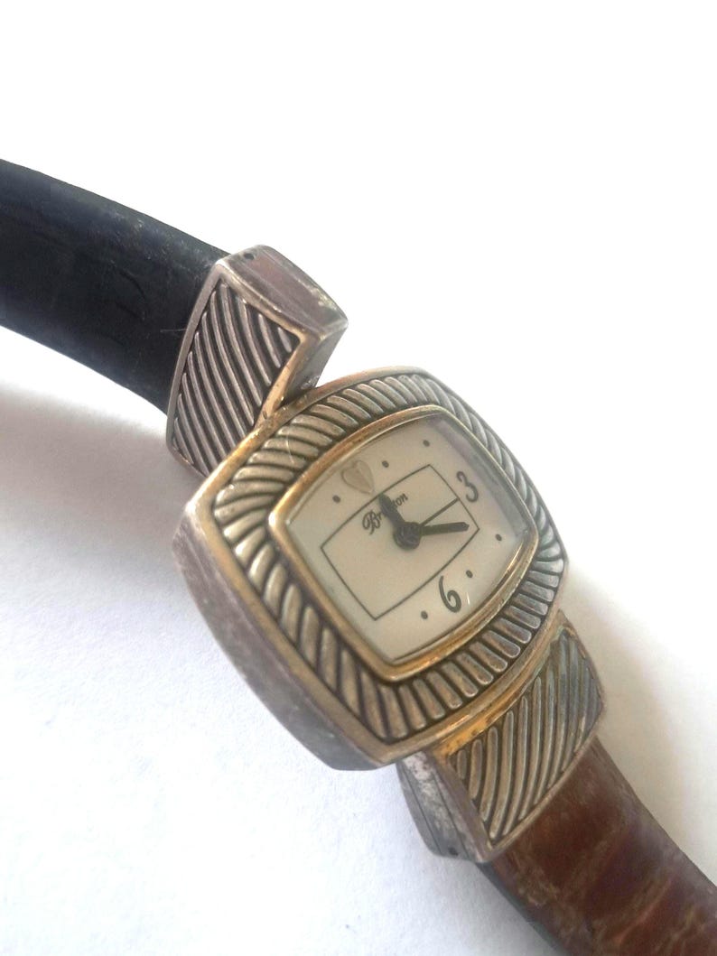 May include: A Brighton watch with a rectangular face and a brown leather band. The watch face is cream-colored with black hands and numerals. The watch has silver-toned metal accents and a black leather strap.