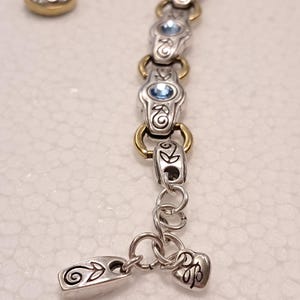 May include: Silver bracelet with gold-coloured accents and light blue gemstones. The bracelet has decorative links and a heart-shaped charm. Two matching stud earrings with blue gemstones are also visible.