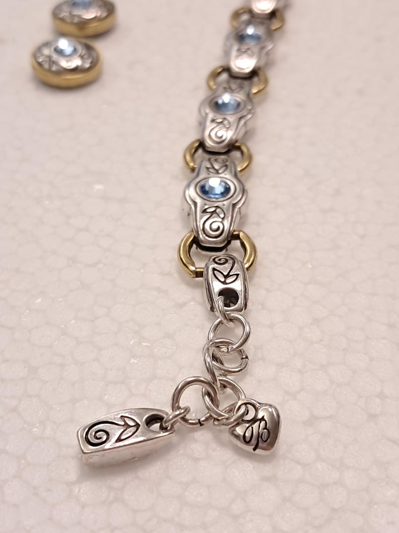 May include: Silver bracelet with gold-coloured accents and light blue gemstones. The bracelet has decorative links and a heart-shaped charm. Two matching stud earrings with blue gemstones are also visible.