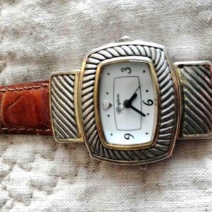 May include: A rectangular Brighton watch with a white face, black hands, and gold-tone accents. The watch has a silver-tone bezel with a braided design and a brown leather band. The watch face displays the numbers 3, 6, and 9.