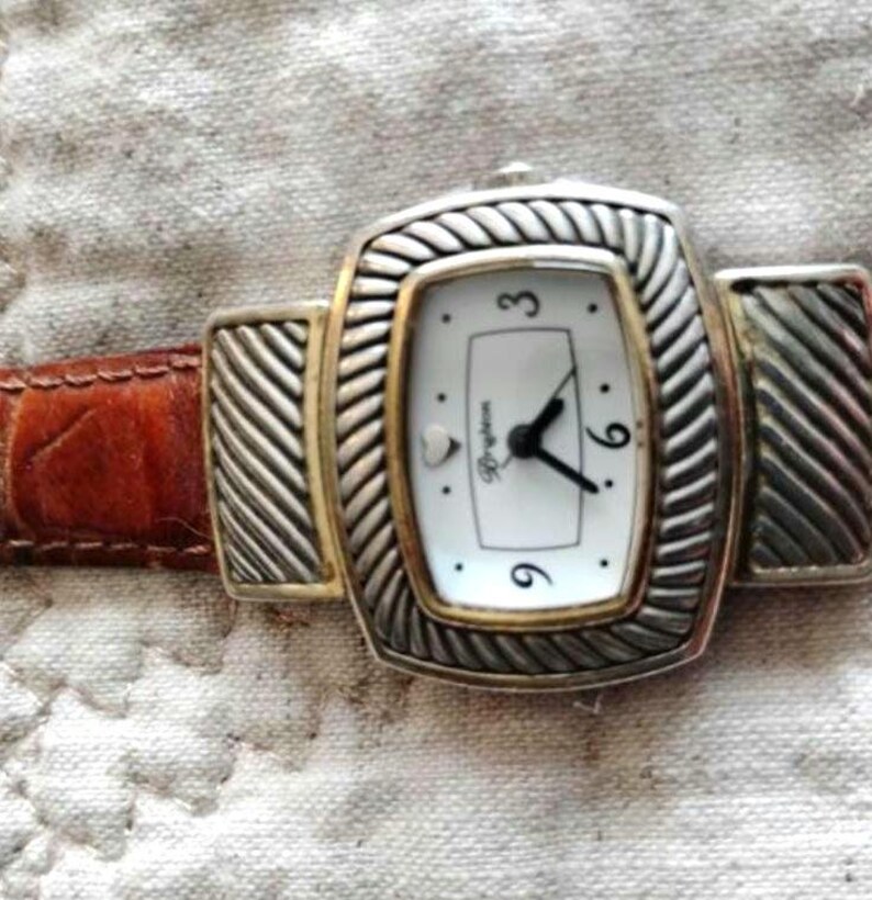 May include: A rectangular Brighton watch with a white face, black hands, and gold-tone accents. The watch has a silver-tone bezel with a braided design and a brown leather band. The watch face displays the numbers 3, 6, and 9.
