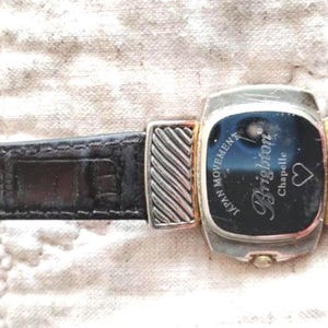 May include: A Brighton Chapelle watch with a black leather band. The watch face is rectangular with a black background and silver and gold accents. The watch has the words "JAPAN MOVEMENT" and "Brighton Chapelle" on the face.