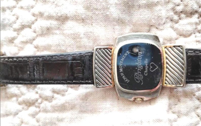 May include: A Brighton Chapelle watch with a black leather band. The watch face is rectangular with a black background and silver and gold accents. The watch has the words "JAPAN MOVEMENT" and "Brighton Chapelle" on the face.