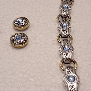 May include: A silver bracelet and matching earrings. The bracelet and earrings have a gold-coloured border and a central light blue gemstone. The design includes decorative swirls and floral accents.