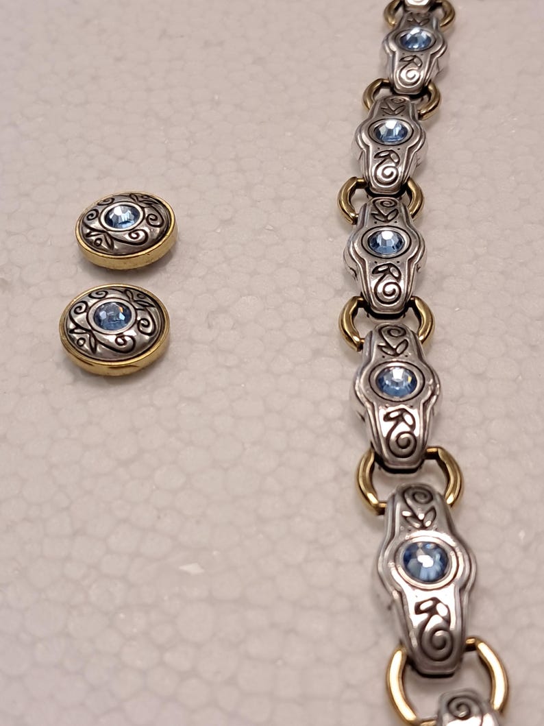 May include: A silver bracelet and matching earrings. The bracelet and earrings have a gold-coloured border and a central light blue gemstone. The design includes decorative swirls and floral accents.