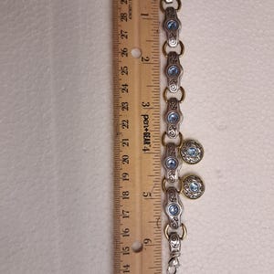 May include: Silver-tone bracelet with gold accents and blue gemstone details. The bracelet is approximately 18 cm long. The design includes oval links with blue stones and round charms with blue centers. A ruler is next to the bracelet.