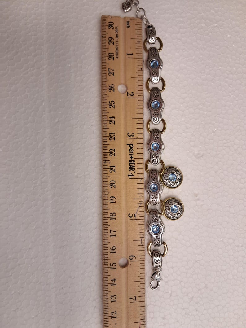 May include: Silver-tone bracelet with gold accents and blue gemstone details. The bracelet is approximately 18 cm long. The design includes oval links with blue stones and round charms with blue centers. A ruler is next to the bracelet.