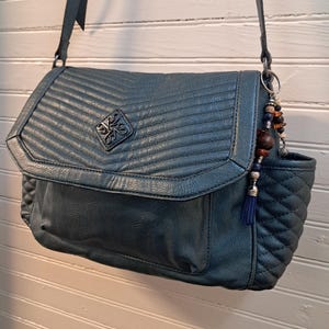 May include: Teal-colored crossbody bag with a flap closure and quilted detailing. The bag features a decorative emblem on the front and a small side pocket. A charm with beads and a tassel hangs from the strap. The bag is made of a textured material.