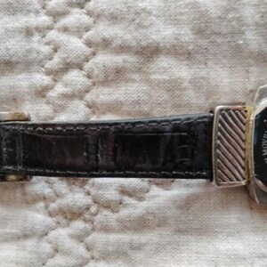 May include: Close-up of a black leather watch strap with a silver buckle and a watch face. The watch face is black with silver accents and the brand name "Brighton". The watch strap is textured.