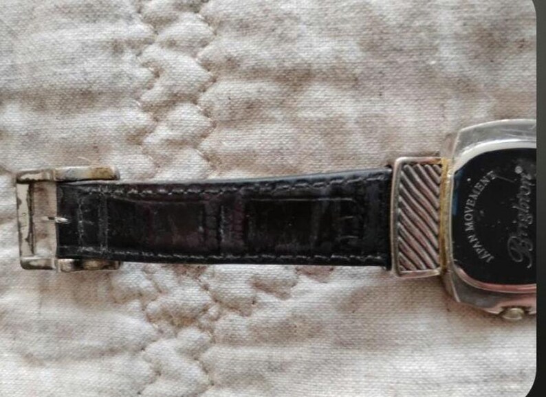May include: Close-up of a black leather watch strap with a silver buckle and a watch face. The watch face is black with silver accents and the brand name "Brighton". The watch strap is textured.