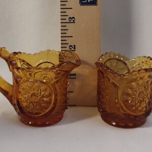 Könnte beinhalten: Ein Set aus zwei Kännchen für Sahne und Zucker aus bernsteinfarbenem Glas mit geschliffenem Glasdesign. Die Kännchen sind klein und haben einen gewellten Rand.
