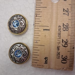 May include: Two round silver and gold-toned buttons with a blue gemstone centre and decorative swirls. The buttons are next to a wooden ruler.