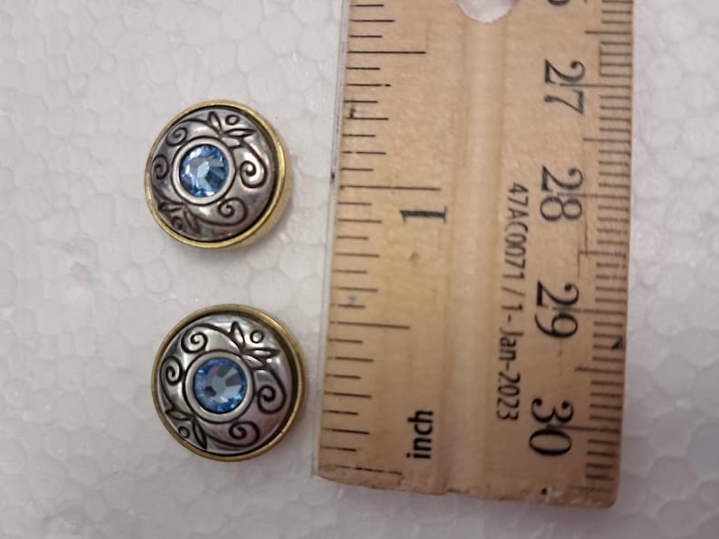 May include: Two round silver and gold-toned buttons with a blue gemstone centre and decorative swirls. The buttons are next to a wooden ruler.