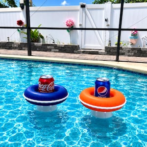 May include: Two circular floating drink holders in a swimming pool. One holder is dark blue with a white stripe, containing a red Coca-Cola can. The other holder is bright orange with a white stripe, containing a blue Pepsi can. Both are buoyant.